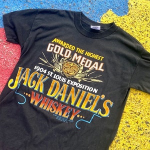 以下が含まれることがあります： ジャックダニエルのウイスキーのヴィンテージロゴがプリントされた黒いTシャツ。ロゴには「Awarded the Highest Gold Medal 1904 St. Louis Exposition Jack Daniel's Whiskey... 」というテキストが黄色とオレンジの文字で書かれています。