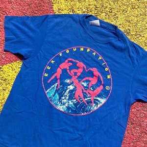 Pode incluir: Uma camiseta azul com um gráfico circular de cinco figuras em rosa dançando em uma cordilheira com um fundo azul e branco. O texto "REX FOUNDATION" está escrito em um círculo ao redor das figuras.