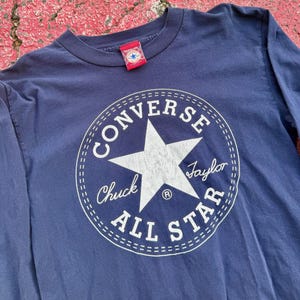May include: Navy blue long-sleeve shirt featuring a large white Converse All Star logo on the front. The logo includes a white star and the words "CONVERSE", "Chuck Taylor", and "ALL STAR". A red tag with a star logo is attached to the collar.