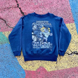 May include: A blue sweatshirt with a graphic of a cartoon mascot wearing a UNC hat and jersey. The text "NORTH CAROLINA TAR HEELS" is printed on the sweatshirt.