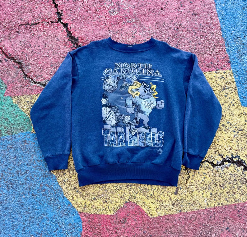 May include: A blue sweatshirt with a graphic of a cartoon mascot wearing a UNC hat and jersey. The text "NORTH CAROLINA TAR HEELS" is printed on the sweatshirt.