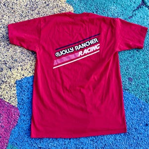 May include: A red t-shirt with the text "@JOLLY RANCHER RACING" in white, blue, and pink. The shirt is a solid color and has short sleeves. The text is on the back of the shirt.