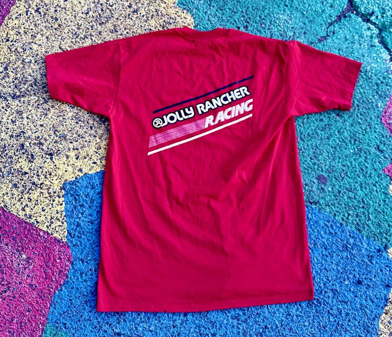 May include: A red t-shirt with the text "@JOLLY RANCHER RACING" in white, blue, and pink. The shirt is a solid color and has short sleeves. The text is on the back of the shirt.
