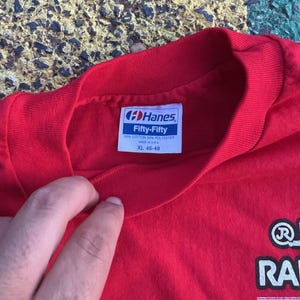 May include: Red Hanes Fifty-Fifty XL t-shirt. The tag reads "50% Cotton 50% Polyester Made in USA". The shirt has a crew neck and a graphic on the lower right side. The shirt is being held by a person.