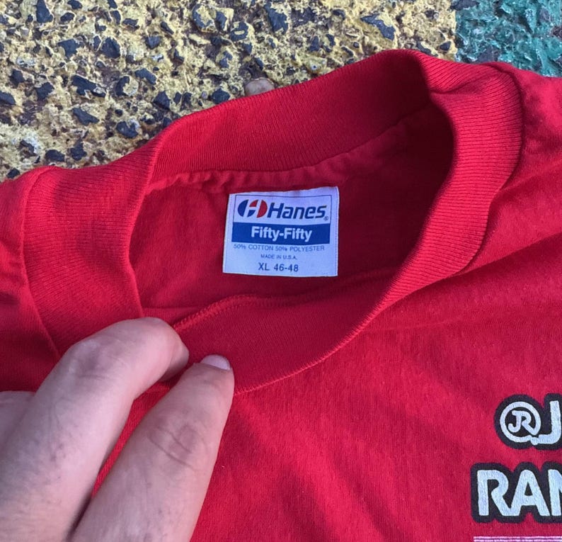 May include: Red Hanes Fifty-Fifty XL t-shirt. The tag reads "50% Cotton 50% Polyester Made in USA". The shirt has a crew neck and a graphic on the lower right side. The shirt is being held by a person.