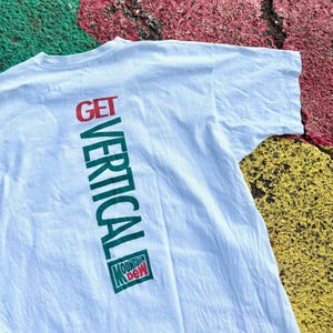 May include: A white short-sleeved t-shirt featuring the text GET VERTICAL printed vertically in red and green ink. Below the text is a green and red Mountain Dew logo. The graphic is positioned on the left side of the shirt fabric.
