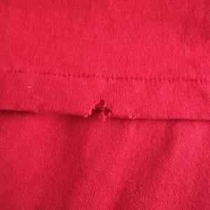 May include: Close-up of a section of red fabric, possibly a t-shirt or other garment. The fabric has a visible knit texture. A small tear is present near the seam, revealing the raw edge of the material.