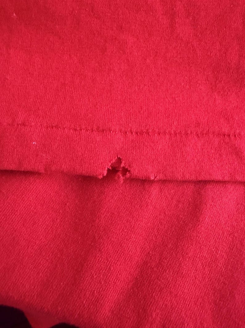 May include: Close-up of a section of red fabric, possibly a t-shirt or other garment. The fabric has a visible knit texture. A small tear is present near the seam, revealing the raw edge of the material.