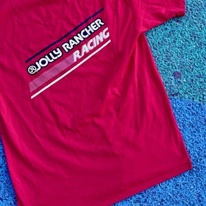 May include: A red t-shirt with the text "@JOLLY RANCHER RACING" in white, blue, and red. The shirt is made of a soft material and has short sleeves. The background is a textured blue and green surface.