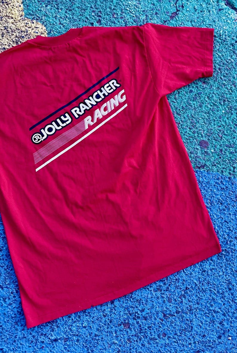 May include: A red t-shirt with the text "@JOLLY RANCHER RACING" in white, blue, and red. The shirt is made of a soft material and has short sleeves. The background is a textured blue and green surface.