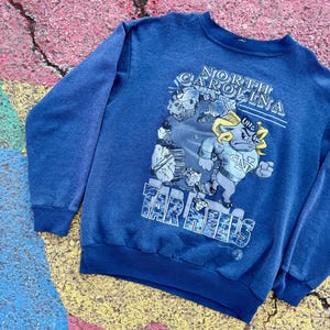 May include: A blue sweatshirt with a graphic print of a cartoon mascot. The mascot is a blue ram with a yellow horn and a white star on its chest. The text "NORTH CAROLINA" and "TAR HEELS" are printed on the sweatshirt.