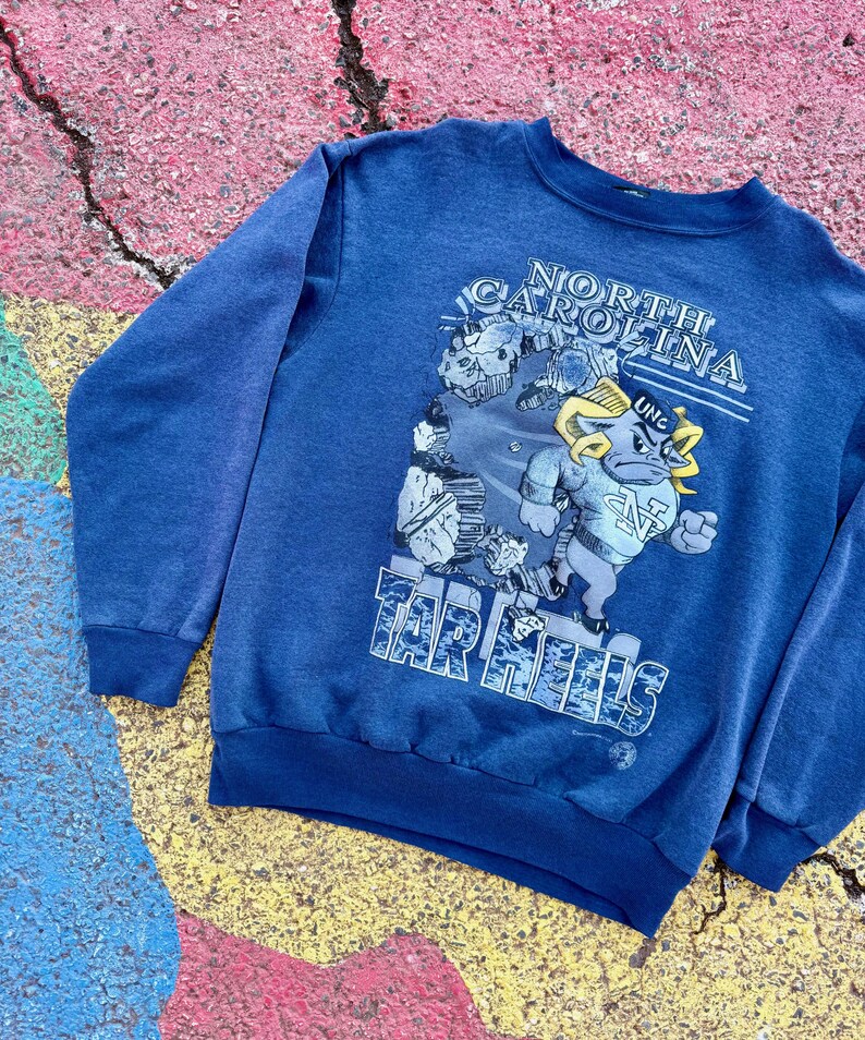 May include: A blue sweatshirt with a graphic print of a cartoon mascot. The mascot is a blue ram with a yellow horn and a white star on its chest. The text "NORTH CAROLINA" and "TAR HEELS" are printed on the sweatshirt.