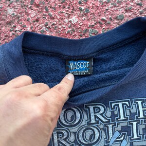 May include: Navy blue sweatshirt with a white graphic print that says "North Carolina" and a small tag that says "Mascot Sportswear Made in U.S.A. L"