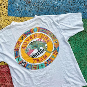 May include: White t-shirt with a colorful graphic on the back. The graphic is a circular design with the words "ADVENTURE TEAM" and "Marlboro" surrounding a lizard illustration. The design includes various geometric shapes and colors.