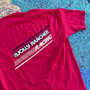 May include: A red t-shirt featuring the text "JOLLY RANCHER RACING" in white and blue diagonal stripes. The shirt is made of a soft material and has a classic crew neck design. The graphic is on the back of the shirt.