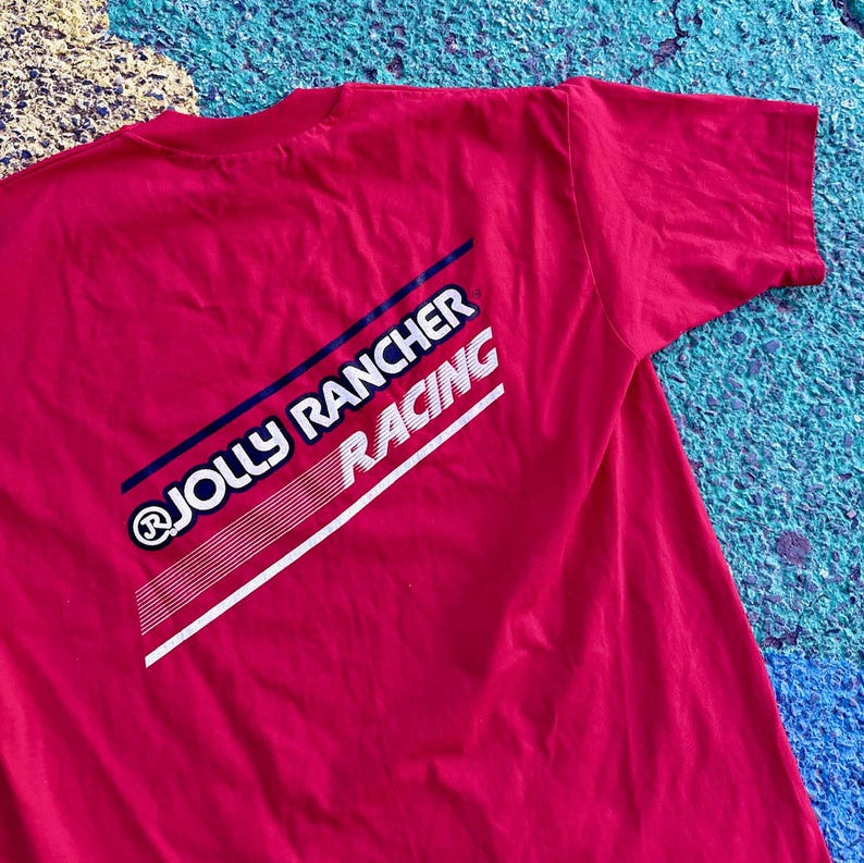 May include: A red t-shirt featuring the text "JOLLY RANCHER RACING" in white and blue diagonal stripes. The shirt is made of a soft material and has a classic crew neck design. The graphic is on the back of the shirt.