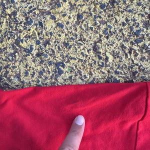 May include: Close-up of a bright red fabric, possibly a shirt or other garment. The fabric is smooth and appears to be made of a soft material. A finger is pointing at the fabric. The background is a textured surface with yellow and black tones.