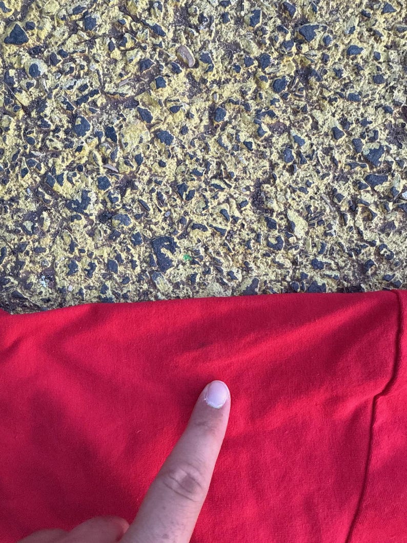 May include: Close-up of a bright red fabric, possibly a shirt or other garment. The fabric is smooth and appears to be made of a soft material. A finger is pointing at the fabric. The background is a textured surface with yellow and black tones.