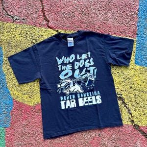 Peut inclure: T-shirt bleu marine avec l'inscription "WHO LET THE DOGS OUT !" et un dessin de chien de dessin anim&eacute;. Le texte "NORTH CAROLINA TAR HEELS" est en dessous. Le t-shirt est pos&eacute; sur une surface craquel&eacute;e multicolore.