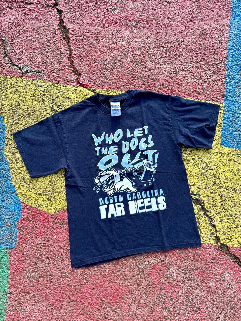 Peut inclure: T-shirt bleu marine avec l'inscription "WHO LET THE DOGS OUT !" et un dessin de chien de dessin anim&eacute;. Le texte "NORTH CAROLINA TAR HEELS" est en dessous. Le t-shirt est pos&eacute; sur une surface craquel&eacute;e multicolore.