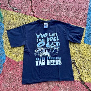 Peut inclure: T-shirt bleu marine avec l'inscription "WHO LET THE DOGS OUT!" en bleu clair. Le t-shirt pr&eacute;sente un dessin de chien de dessin anim&eacute; et les mots "NORTH CAROLINA TAR HEELS" en blanc.