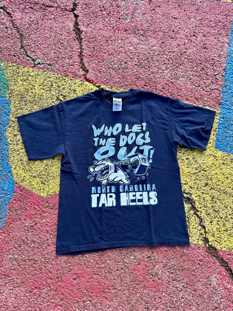 Peut inclure: T-shirt bleu marine avec l'inscription "WHO LET THE DOGS OUT!" en bleu clair. Le t-shirt pr&eacute;sente un dessin de chien de dessin anim&eacute; et les mots "NORTH CAROLINA TAR HEELS" en blanc.