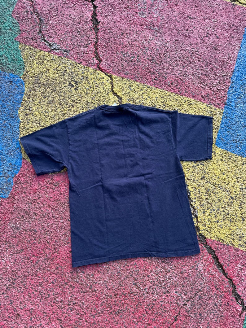 Peut inclure: Un t-shirt bleu marine &agrave; manches courtes, pos&eacute; &agrave; plat. Le t-shirt est fait d'une mati&egrave;re douce. Le fond est une surface color&eacute;e et textur&eacute;e avec des taches roses, jaunes, bleues et vertes.