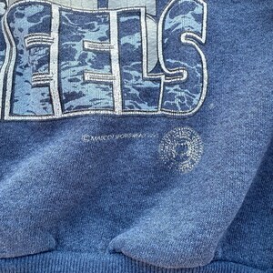 May include: A blue sweatshirt with a faded graphic of the word "FEELS" in a camouflage pattern. The sweatshirt has a ribbed hem and a small copyright symbol with the text "MARV ORISHREDZ 2004".