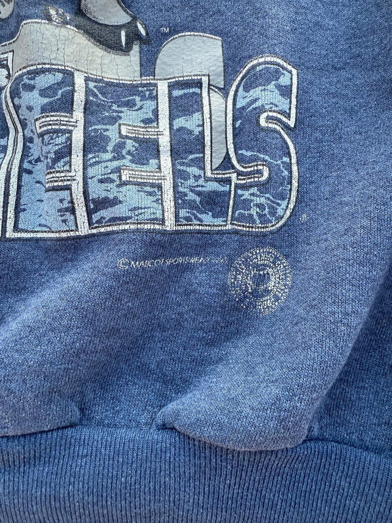 May include: A blue sweatshirt with a faded graphic of the word "FEELS" in a camouflage pattern. The sweatshirt has a ribbed hem and a small copyright symbol with the text "MARV ORISHREDZ 2004".
