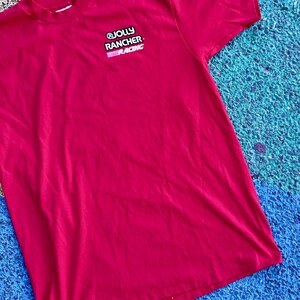 May include: A red t-shirt with the "JOLLY RANCHER RACING" logo on the upper left chest. The logo is in white and pink. The shirt is made of a soft material and is laying on a blue and green surface.