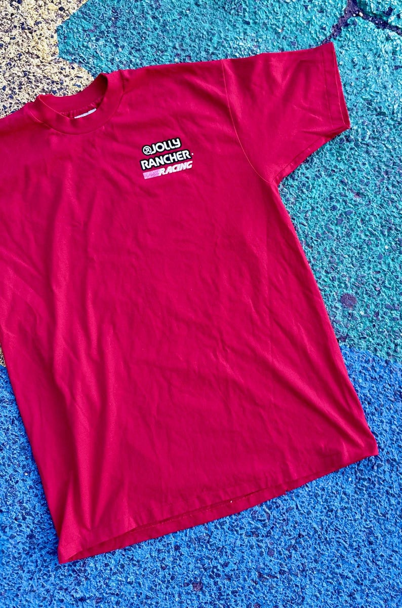 May include: A red t-shirt with the "JOLLY RANCHER RACING" logo on the upper left chest. The logo is in white and pink. The shirt is made of a soft material and is laying on a blue and green surface.