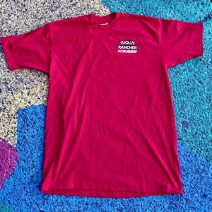 May include: A short-sleeved, magenta-colored t-shirt. The shirt features the Jolly Rancher Racing logo on the upper left chest. The logo is white, black, and pink. The shirt is made of a soft material.