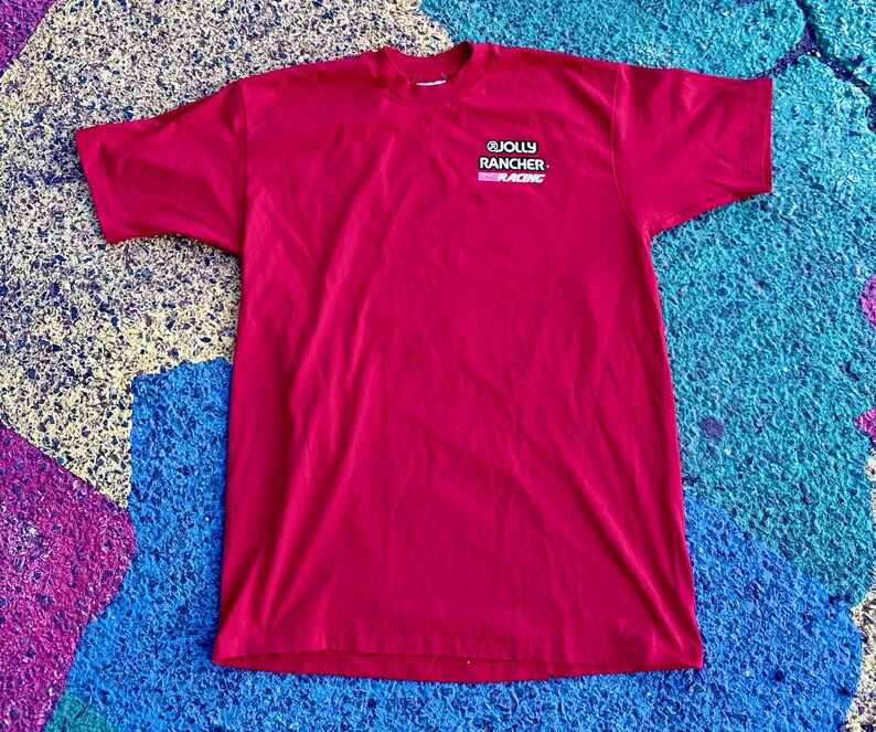 May include: A short-sleeved, magenta-colored t-shirt. The shirt features the Jolly Rancher Racing logo on the upper left chest. The logo is white, black, and pink. The shirt is made of a soft material.