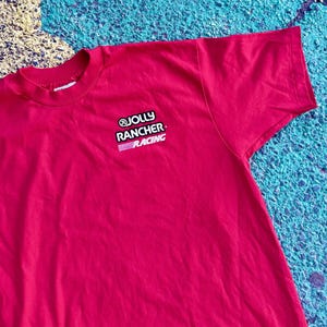 May include: A red t-shirt with the text "@JOLLY RANCHER RACING" in white and pink. The shirt is made of a soft material and has a crew neck. The t-shirt is a solid red color.
