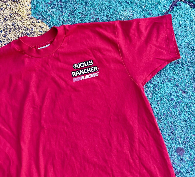 May include: A red t-shirt with the text "@JOLLY RANCHER RACING" in white and pink. The shirt is made of a soft material and has a crew neck. The t-shirt is a solid red color.