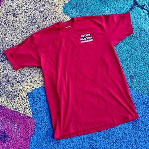 May include: A red t-shirt with the text "JOLLY RANCHER RACING" printed on the upper left chest. The shirt is made of a soft material and is laying on a colorful surface.