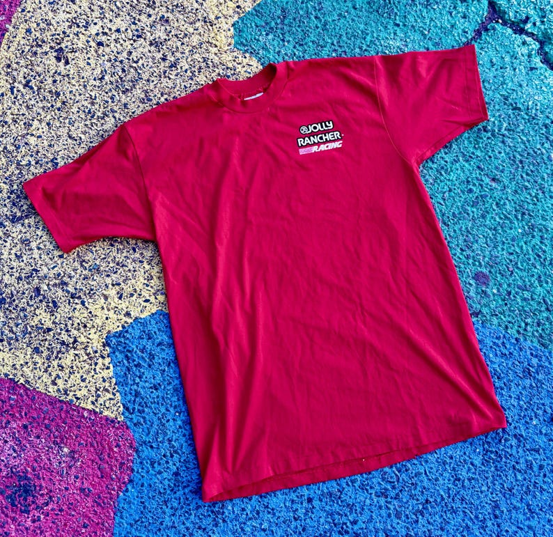May include: A red t-shirt with the text "JOLLY RANCHER RACING" printed on the upper left chest. The shirt is made of a soft material and is laying on a colorful surface.