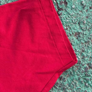 May include: Close-up of a section of a bright red t-shirt. The fabric is smooth and appears to be a knit material. The edge of the shirt is visible, showing a neatly sewn hem. The background is a textured, mottled green surface.