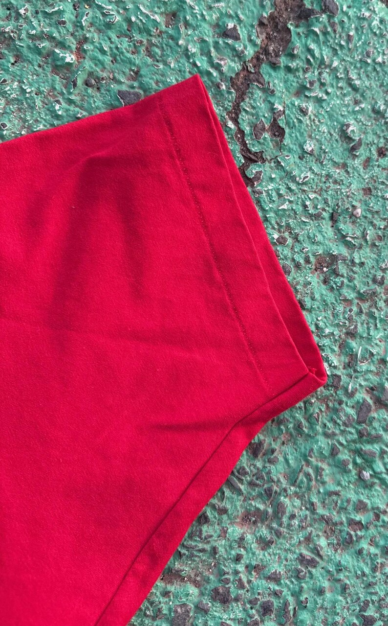 May include: Close-up of a section of a bright red t-shirt. The fabric is smooth and appears to be a knit material. The edge of the shirt is visible, showing a neatly sewn hem. The background is a textured, mottled green surface.