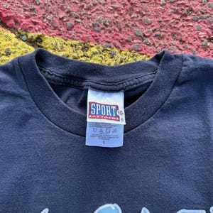 Peut inclure: T-shirt bleu marine avec un motif graphique bleu clair. Le t-shirt a un col rond et une &eacute;tiquette qui indique "SPORT ATTACK". Le t-shirt est de taille L.