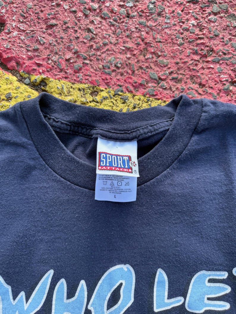 Peut inclure: T-shirt bleu marine avec un motif graphique bleu clair. Le t-shirt a un col rond et une &eacute;tiquette qui indique "SPORT ATTACK". Le t-shirt est de taille L.