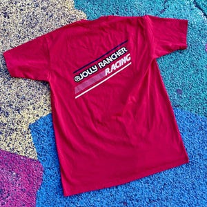May include: A red t-shirt with the text "JOLLY RANCHER RACING" printed on the back in white, navy blue, and red. The shirt is laid flat on a multi-colored surface.