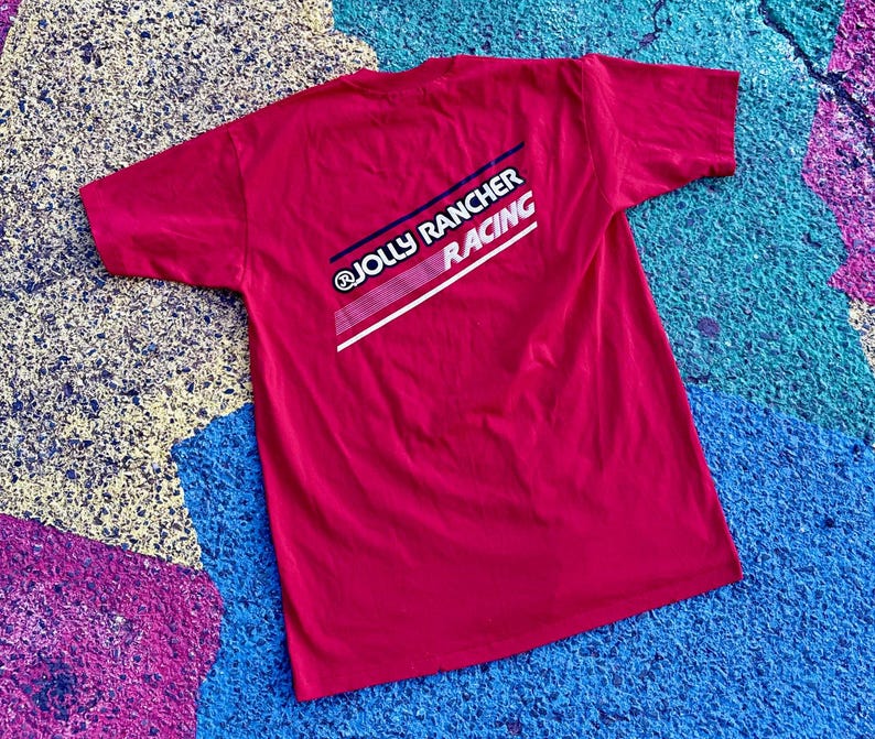 May include: A red t-shirt with the text "JOLLY RANCHER RACING" printed on the back in white, navy blue, and red. The shirt is laid flat on a multi-colored surface.