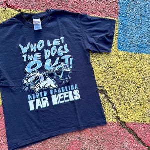 Peut inclure: T-shirt bleu marine avec l'inscription "WHO LET THE DOGS OUT!" en bleu clair et blanc. Le t-shirt pr&eacute;sente un dessin de chien de dessin anim&eacute; et les mots "NORTH CAROLINA TAR HEELS" en blanc.