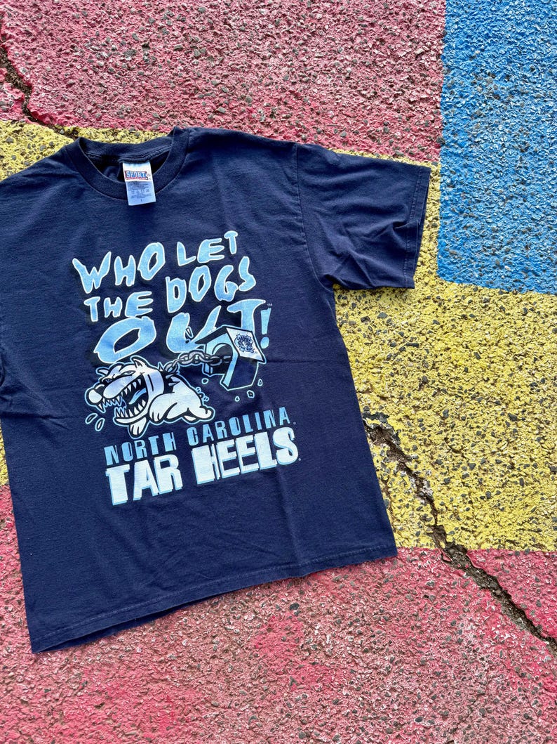 Peut inclure: T-shirt bleu marine avec l'inscription "WHO LET THE DOGS OUT!" en bleu clair et blanc. Le t-shirt pr&eacute;sente un dessin de chien de dessin anim&eacute; et les mots "NORTH CAROLINA TAR HEELS" en blanc.