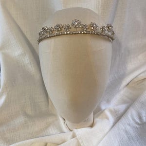 Pode incluir: Uma tiara prateada com flores de strass transparentes. A tiara está em uma cabeça de manequim branca.