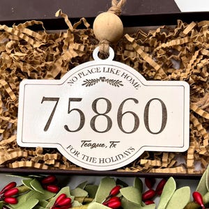Könnte beinhalten: Cremefarbener Weihnachtsschmuck mit dem Text "NO PLACE LIKE HOME" und "75860 Teague, TX FOR THE HOLIDAYS". Der Anhänger ist rechteckig mit dekorativem Rand und einer Holzperle oben.