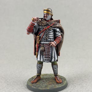 Pode incluir: Uma figura em miniatura de um soldado romano, pintada com armadura detalhada, uma capa castanha e um capacete. A figura segura uma espada e está sobre uma pequena base texturizada. A paleta de cores inclui prata, vermelho, castanho e ouro.