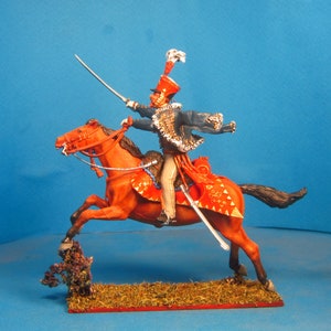 May include: A hand-painted miniature figure of a mounted soldier in a blue uniform with a red sash and a white plume on his hat. The soldier is riding a red horse with gold trim and is holding a sword.