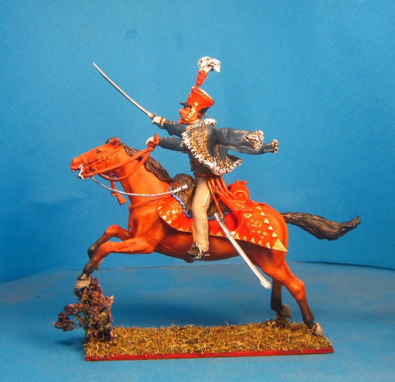 May include: A hand-painted miniature figure of a mounted soldier in a blue uniform with a red sash and a white plume on his hat. The soldier is riding a red horse with gold trim and is holding a sword.